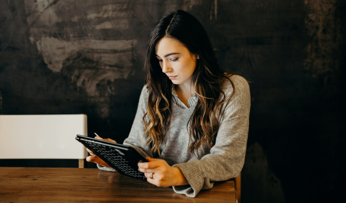 Woman at a wooden table using a tablet and stylus to work through the costs of a business idea